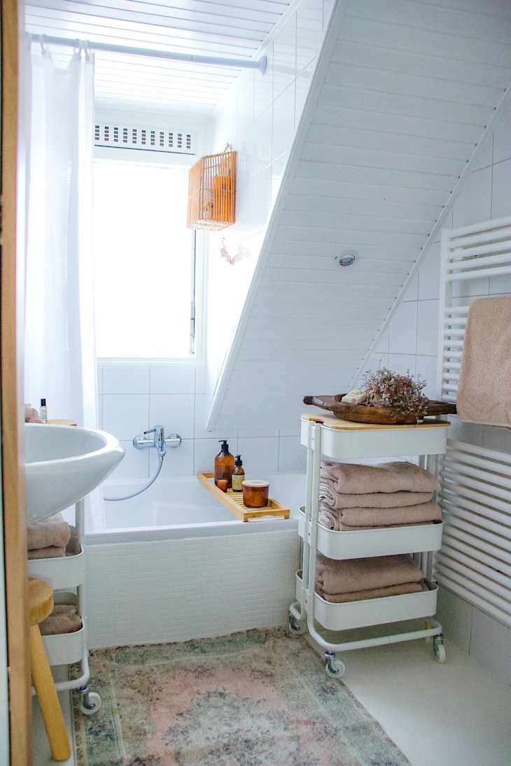 Bright bathroom with a bathtub, sink, wooden towel rack, and a cart holding towels and toiletries.
