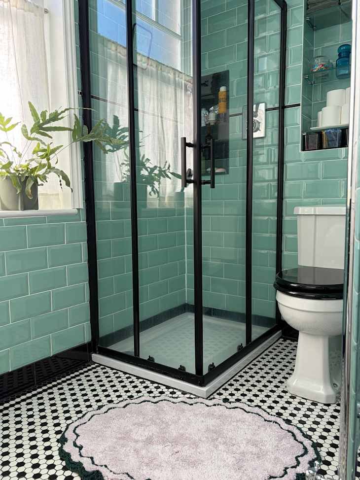 Modern bathroom featuring a glass shower, teal subway tiles, black and white patterned floor, and a plush lavender rug.
